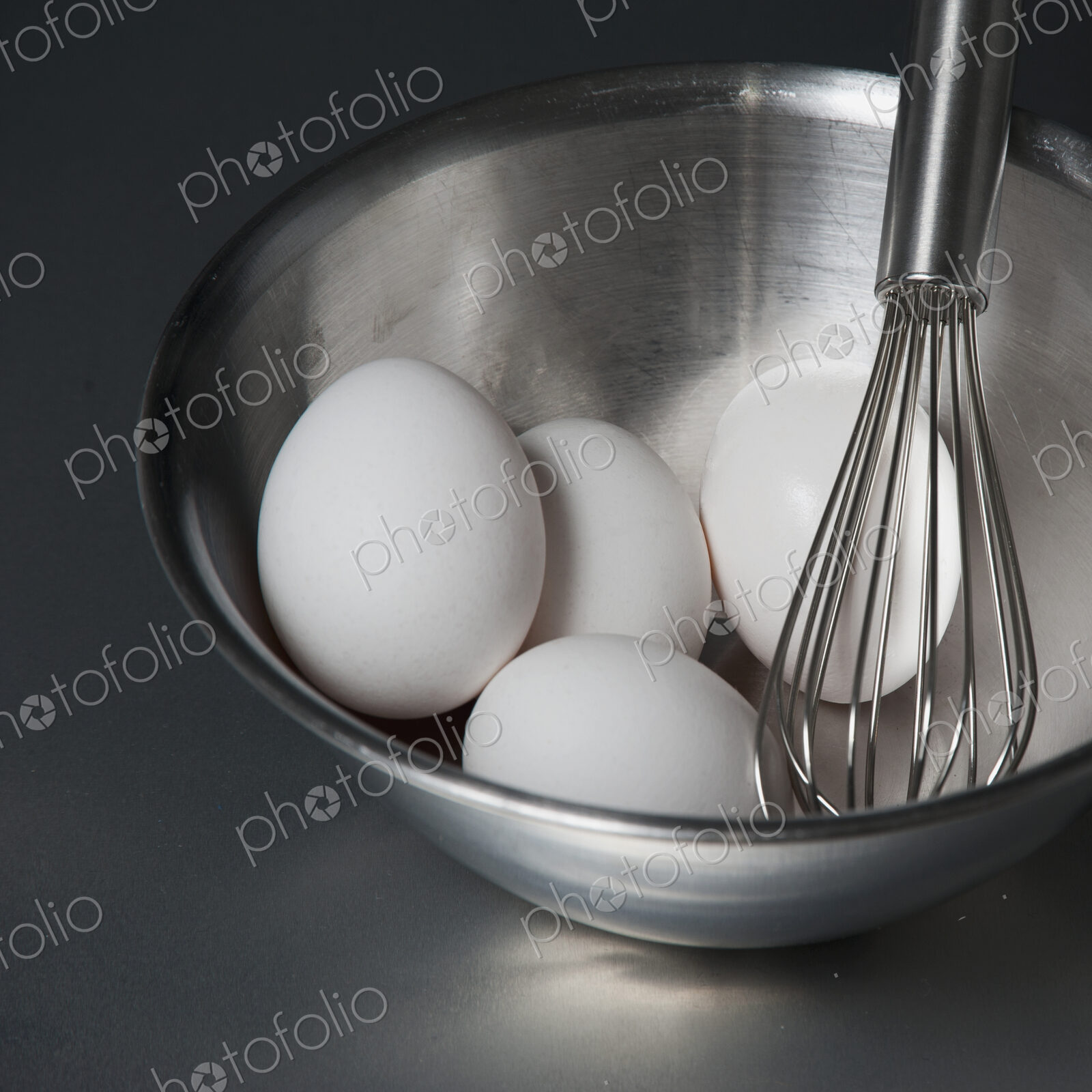 A metal bowl with a balloon whisk and white eggs.