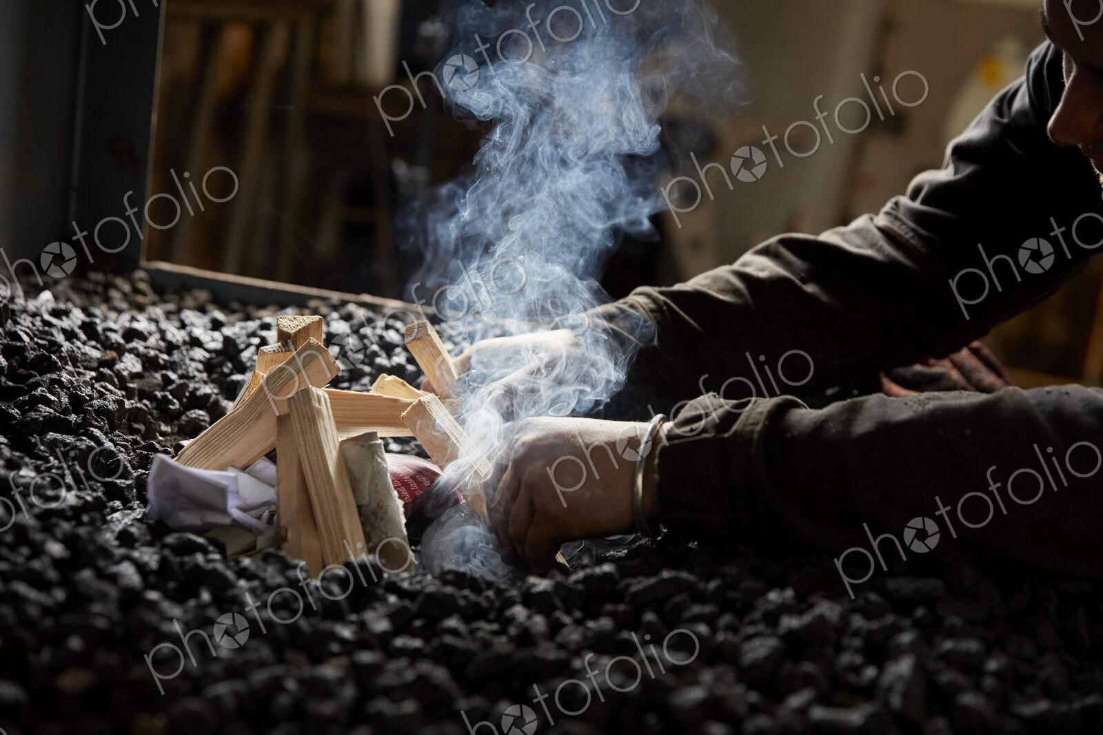 A metal worker lighting a furnace fire using wooden sticks and paper on top of a heap…