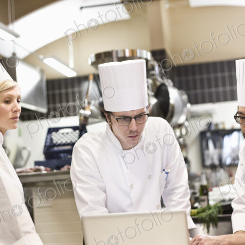 A mixed race group of chefs working with a laptop computer in a commercial kitchen. A mixed race group of chefs working with a laptop computer in a commercial kitchen.