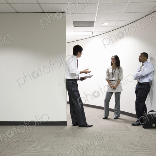 A mixed race group of three businesspeople standing and talking in an office hallway.