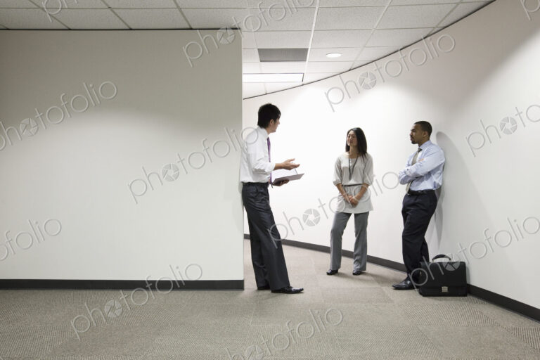A mixed race group of three businesspeople standing and talking in an office hallway.