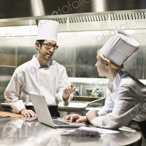 A mixed race pair of chefs working with a laptop computer in a commercial kitchen. A mixed race pair of chefs working with a laptop computer in a commercial kitchen.