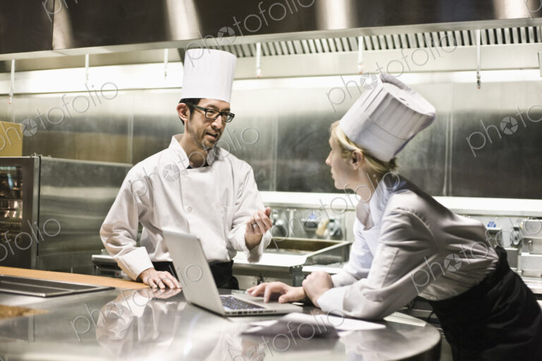 A mixed race pair of chefs working with a laptop computer in a commercial kitchen.