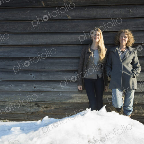 A mother and daughter standing leaning against the wooden wall of a barn on a farm.…