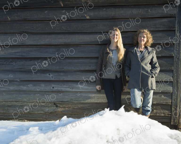 A mother and daughter standing leaning against the wooden wall of a barn on a farm.…