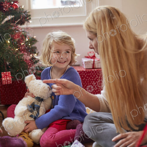 A mother and daughter unwrapping presents under a Christmas tree, holding a teddy bear.