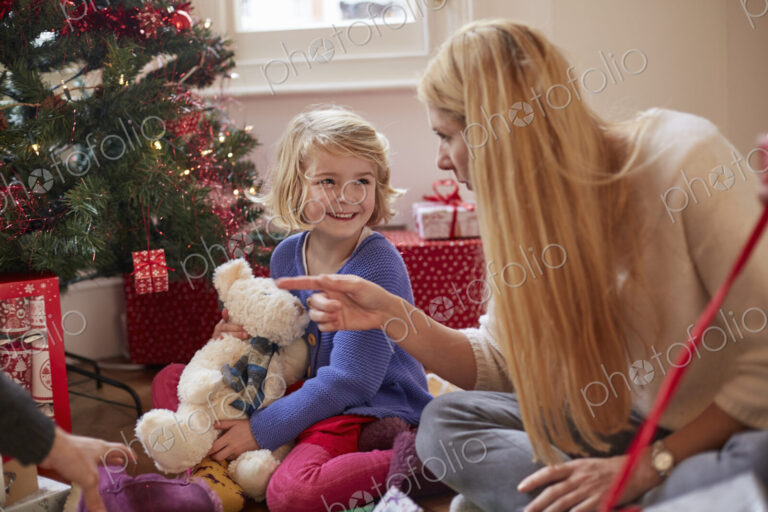 A mother and daughter unwrapping presents under a Christmas tree, holding a teddy bear.