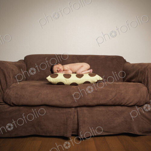 A naked newborn baby lying on his front, sleeping on a pillow on a brown couch. A naked newborn baby lying on his front, sleeping on a pillow on a brown couch.