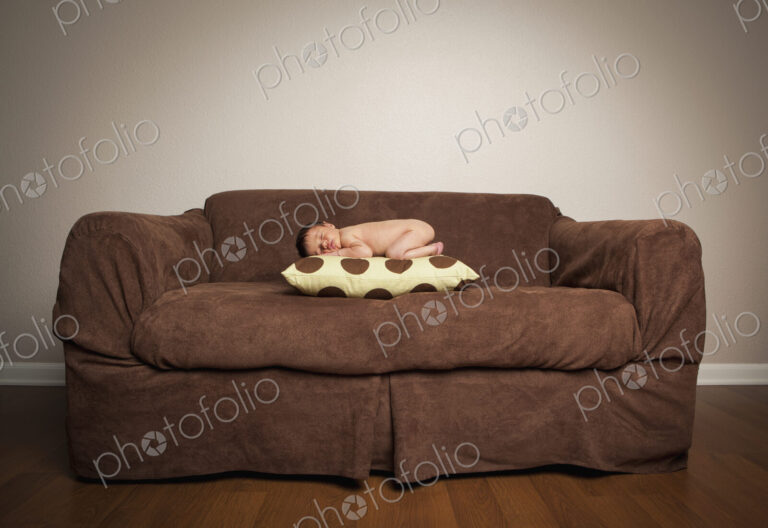A naked newborn baby lying on his front, sleeping on a pillow on a brown couch.