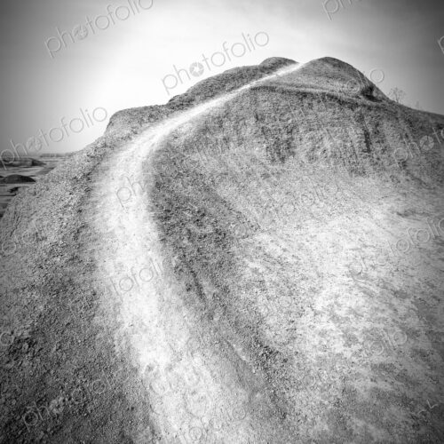 A narrow dirt trail curves up a barren hill under a bright sky in Badlands National… A narrow dirt trail curves up a barren hill under a bright sky in Badlands National…