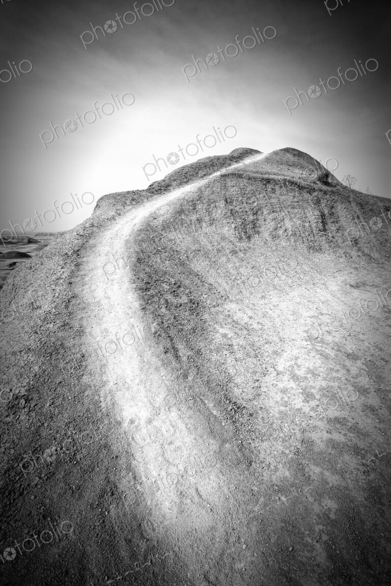 A narrow dirt trail curves up a barren hill under a bright sky in Badlands National…
