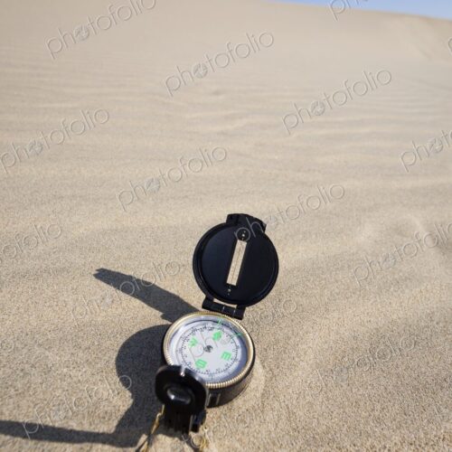 A navigation compass rests on a sandy dune beneath a vibrant blue sky, representing…