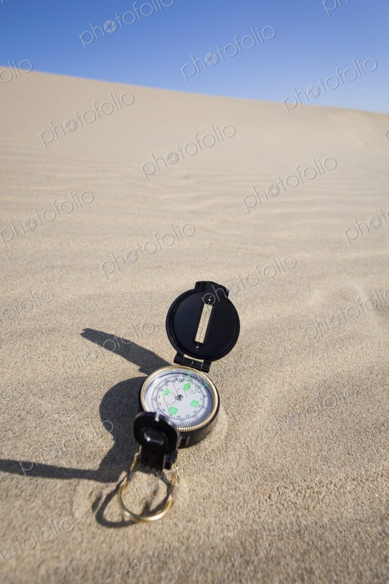 A navigation compass rests on a sandy dune beneath a vibrant blue sky, representing…