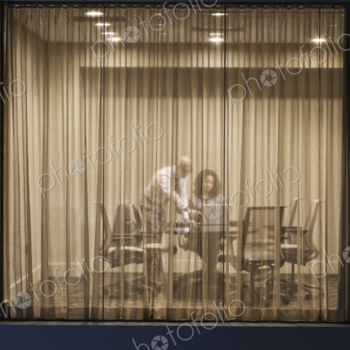 A night view looking into a conference room with two businesspeople working at… A night view looking into a conference room with two businesspeople working at…