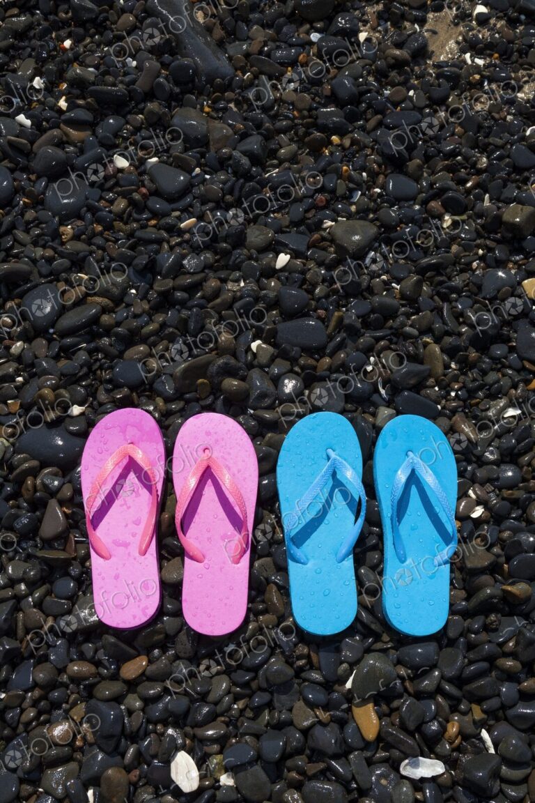 A pair of pink flip flops and a pair of blue flip flops lie side-by-side on a rocky…