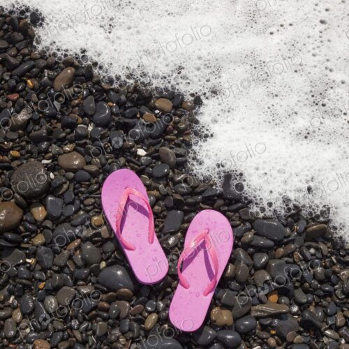 A pair of pink flip flops rest on wet rocks as a foamy ocean wave approaches the shore… A pair of pink flip flops rest on wet rocks as a foamy ocean wave approaches the shore…