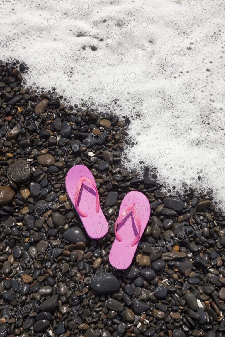 A pair of pink flip flops rest on wet rocks as a foamy ocean wave approaches the shore…