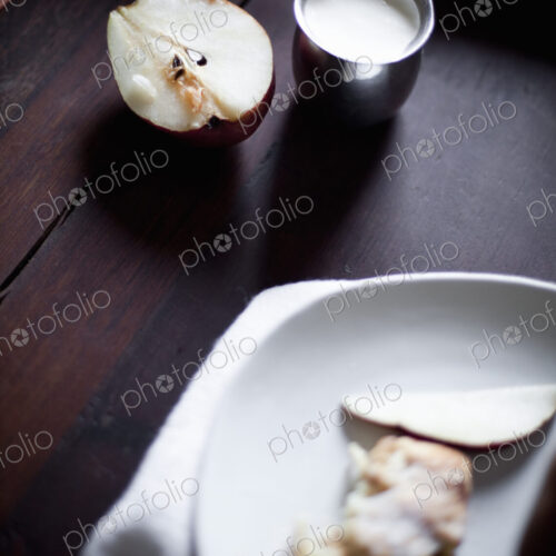 A pear fruit cut open, and sliced. Served on a white china plate. A jug of cream. A…