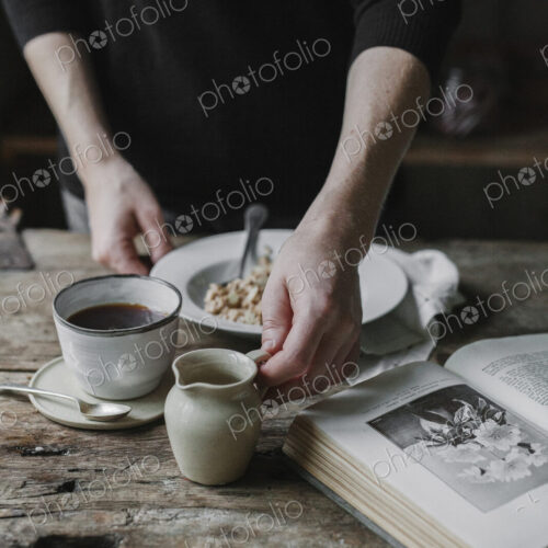 A person at a table with a cup of coffee, bowl of muesli and an open book.