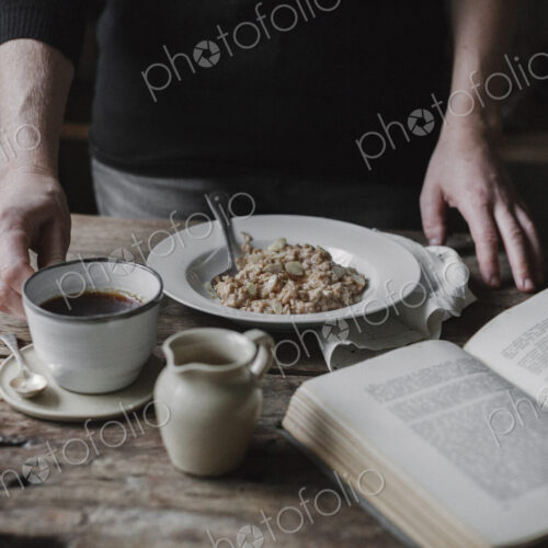A person at a table with a cup of coffee, bowl of muesli and an open book.
