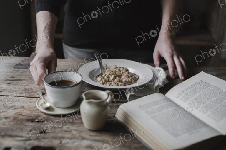 A person at a table with a cup of coffee, bowl of muesli and an open book.