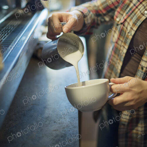 A person, barista, making coffee, and pouring frothed milk from a jug into a cup for a…