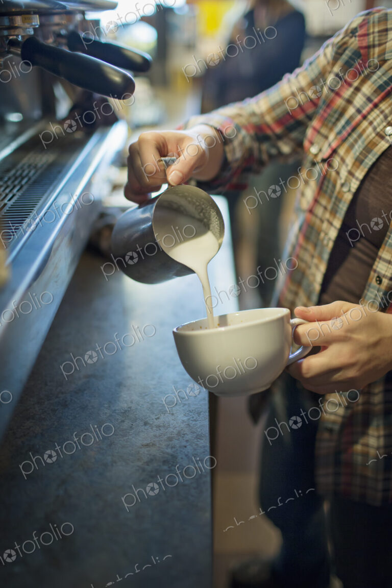 A person, barista, making coffee, and pouring frothed milk from a jug into a cup for a…