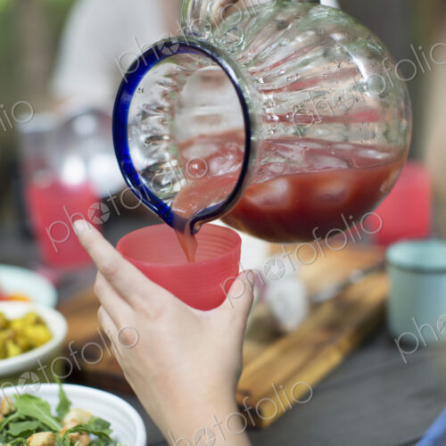 A person holding a cup and pouring iced tomato juice from a large jug. A person holding a cup and pouring iced tomato juice from a large jug.