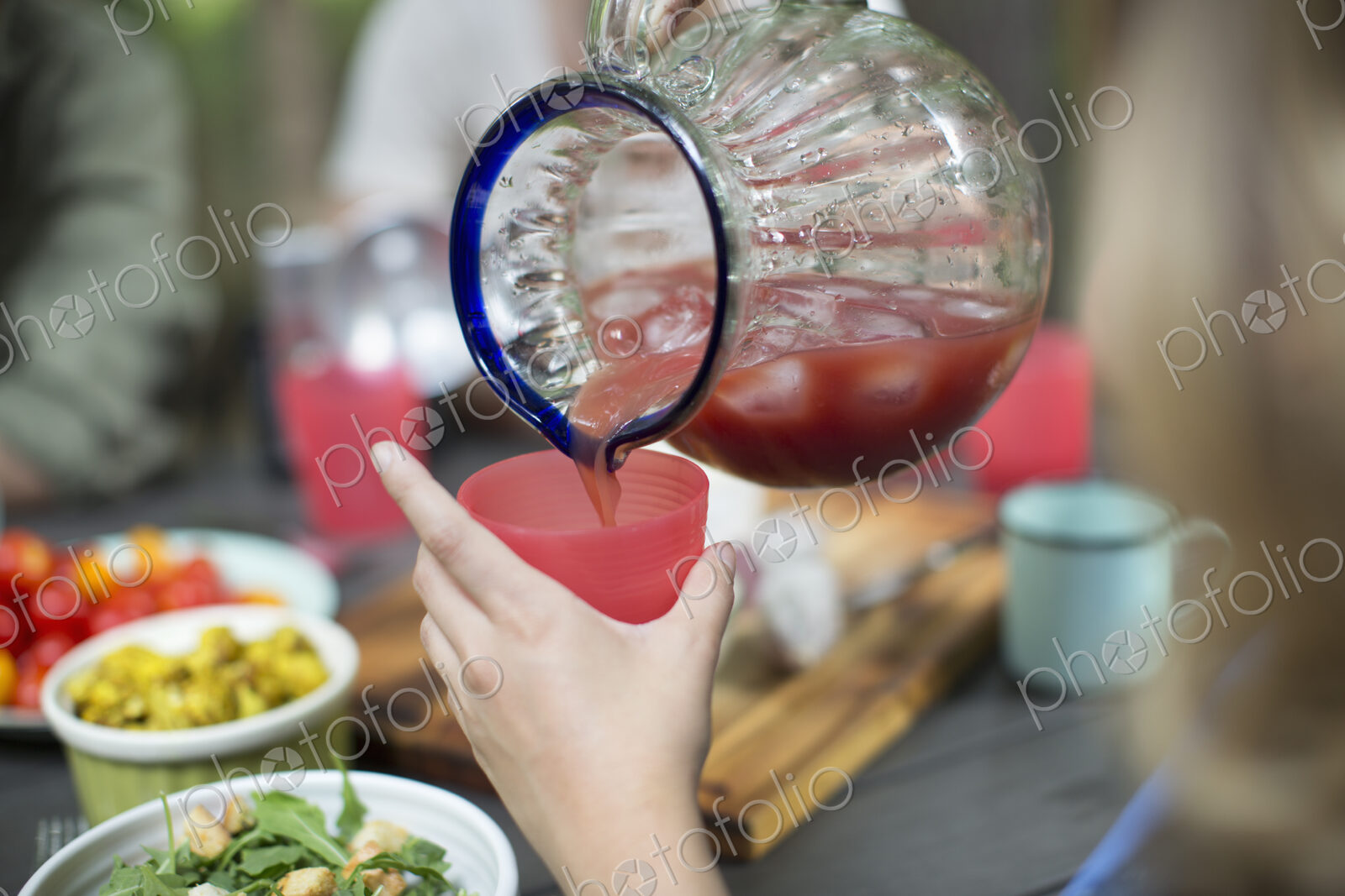 A person holding a cup and pouring iced tomato juice from a large jug.