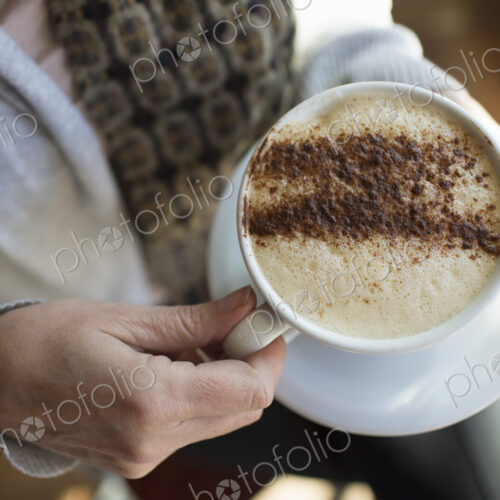 A person holding a full cup of frothy cappuccino coffee in a white china cup.…