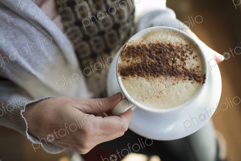 A person holding a full cup of frothy cappuccino coffee in a white china cup.…