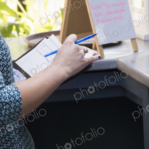 A person in a cafe holding a bill and a pen standing by a bar counter, a menu board on… A person in a cafe holding a bill and a pen standing by a bar counter, a menu board on…