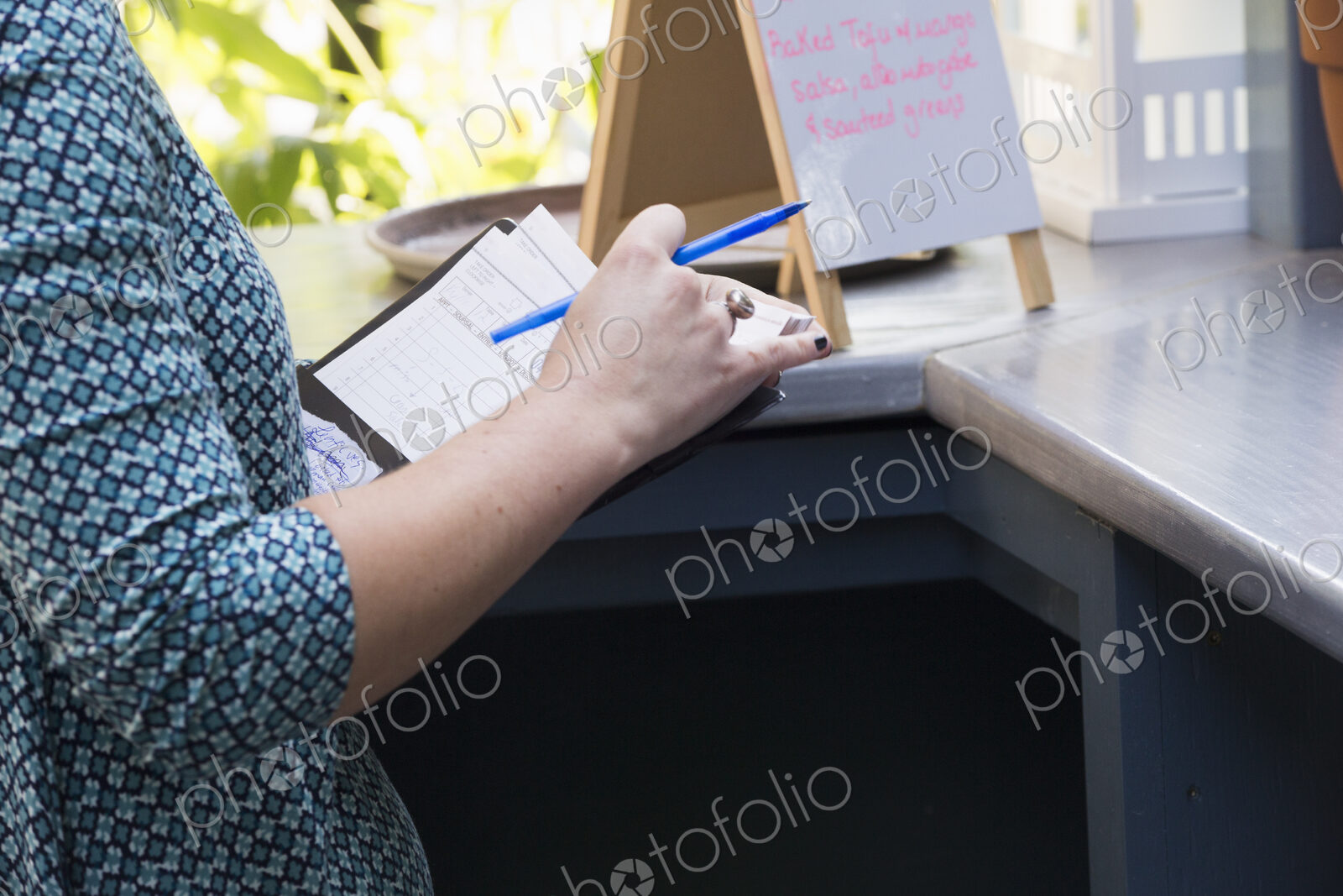 A person in a cafe holding a bill and a pen standing by a bar counter, a menu board on…