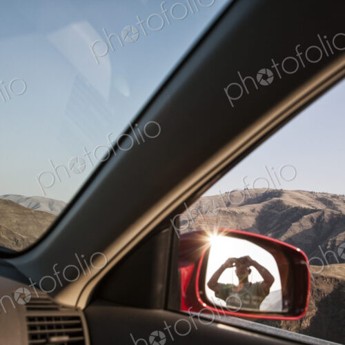 A person lookng through binoculars reflected in the side mirror of a car at a rest stop.