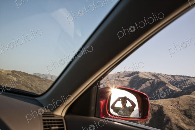 A person lookng through binoculars reflected in the side mirror of a car at a rest stop.