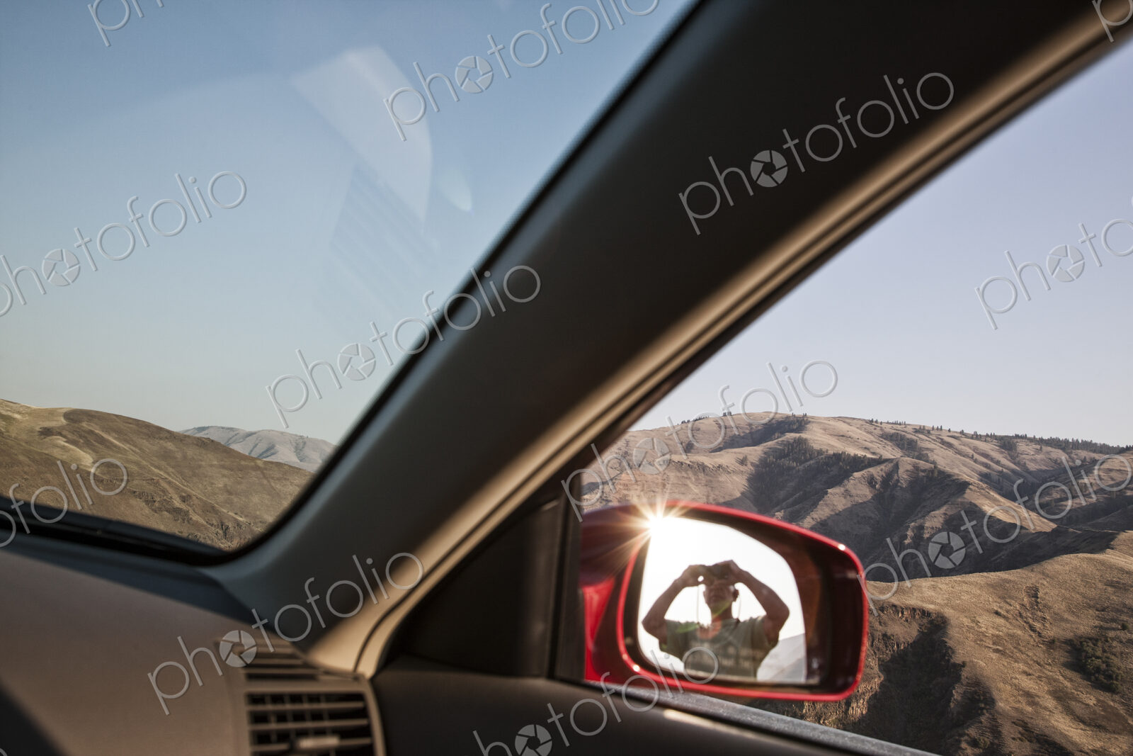 A person lookng through binoculars reflected in the side mirror of a car at a rest stop.