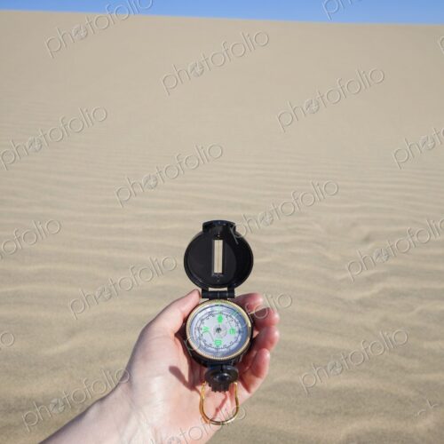 A persons hand holding a navigation compass against the backdrop of a sandy dune under…