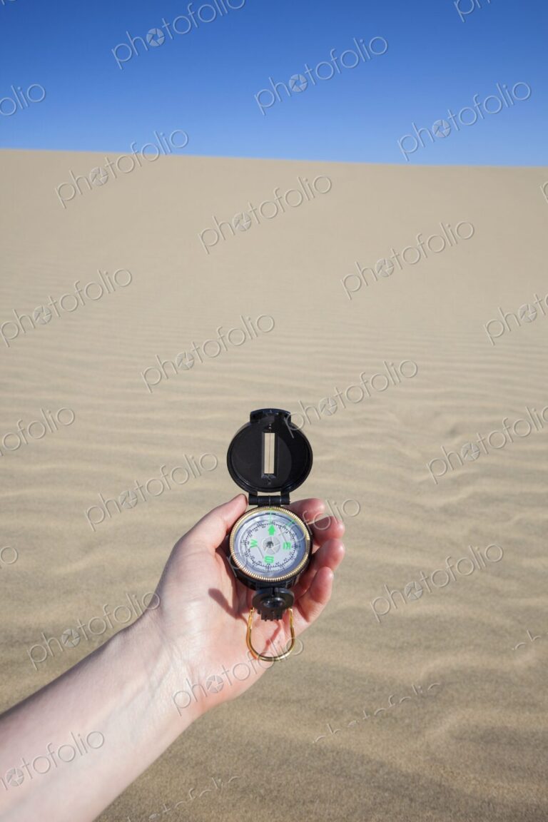 A persons hand holding a navigation compass against the backdrop of a sandy dune under…