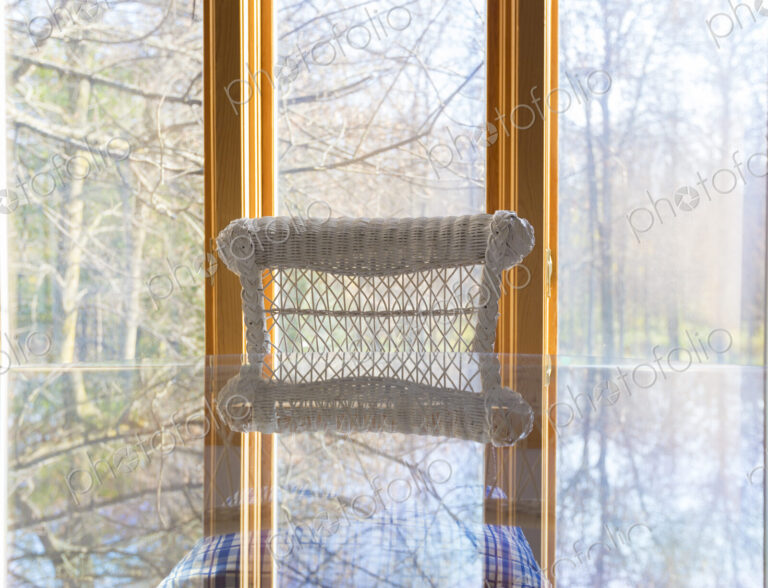 A picture window view to woodland, a glass table and white cane chair.