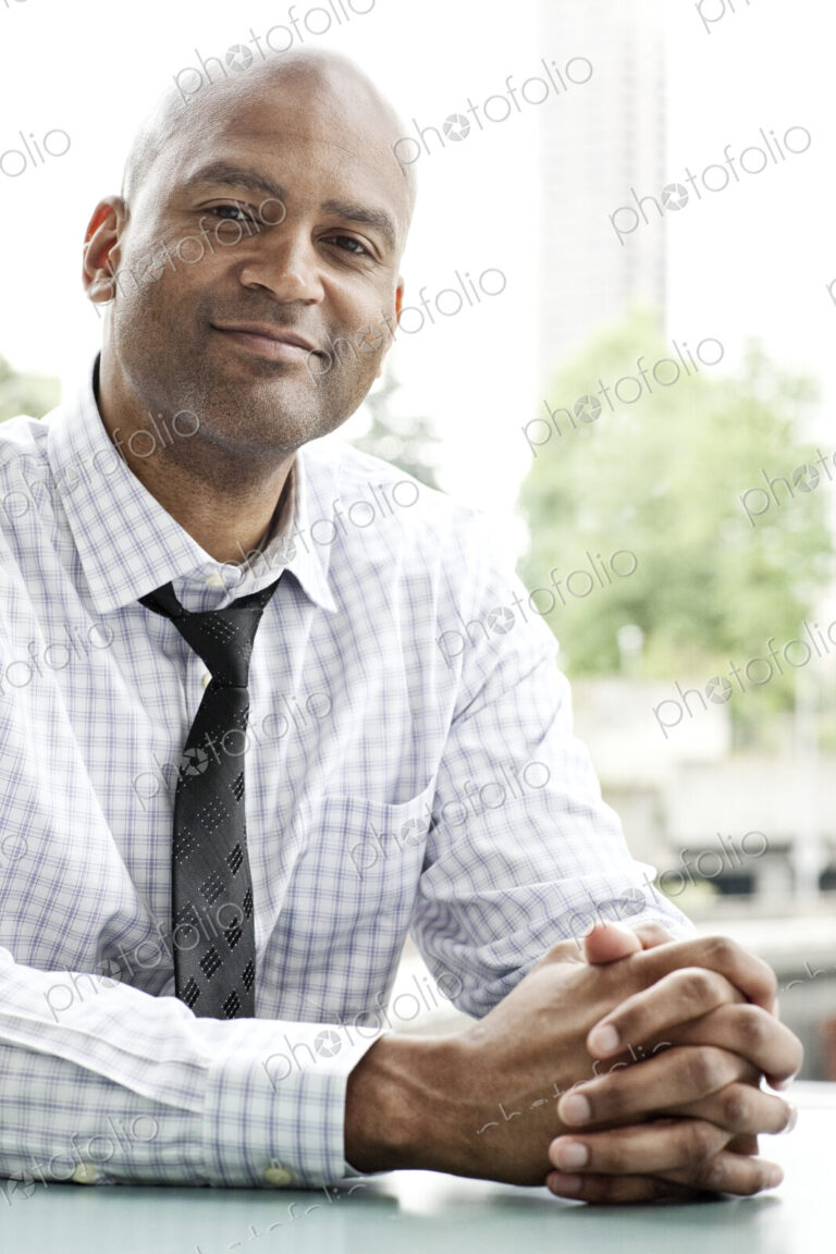 A portrait of a black businessman in a shirt and tie sitting at a desk next to a window.