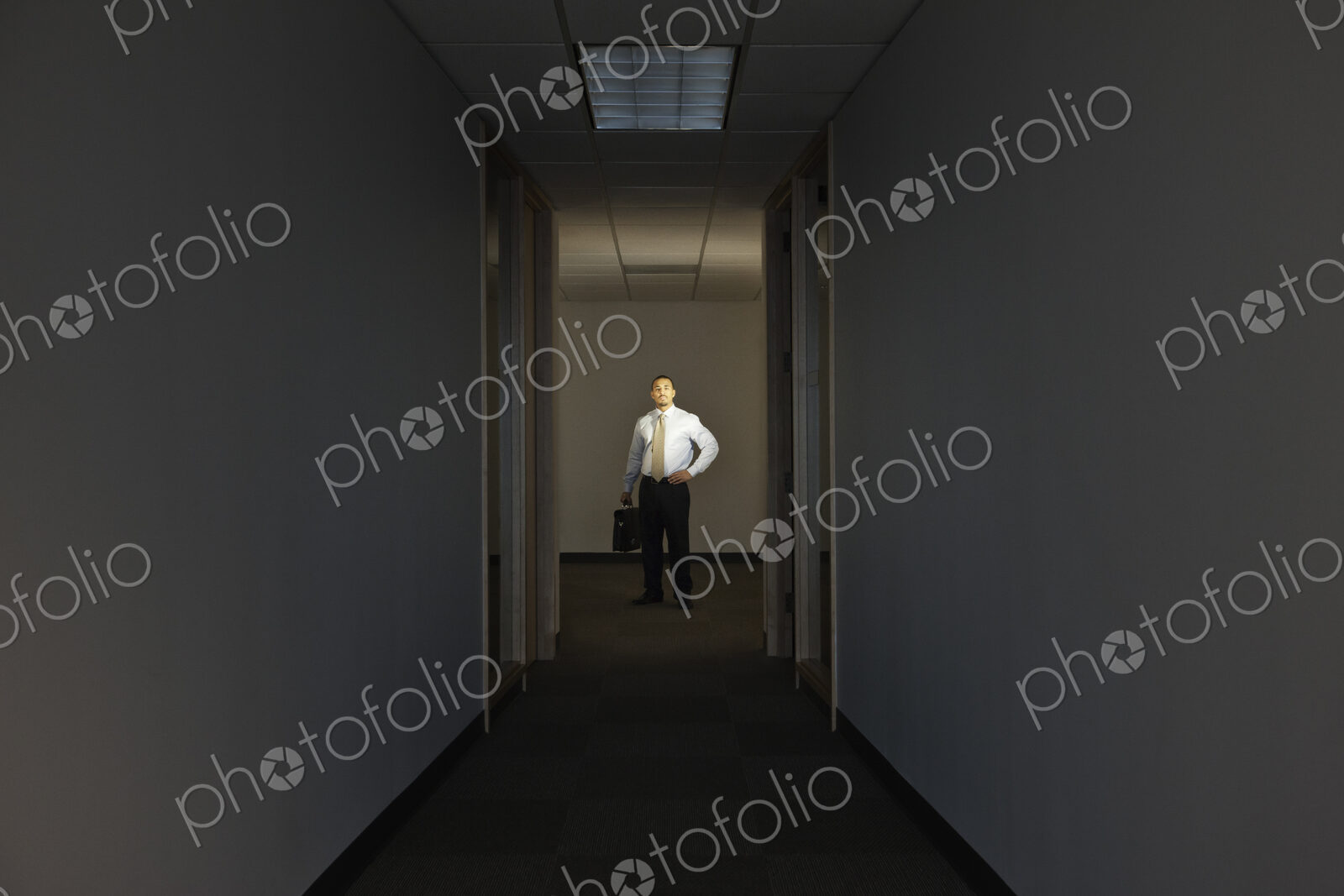 A portrait of a black businessman standing at the end of a long dark hallway.