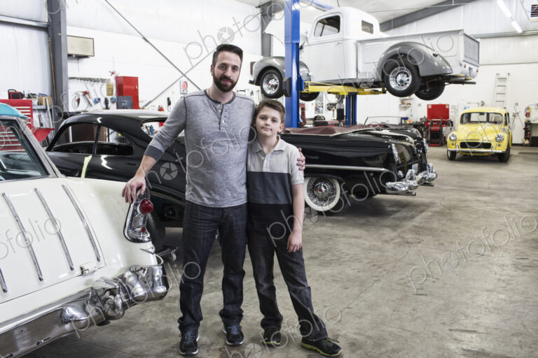 A portrait of a caucasian male and his young son in their classic car repair shop.