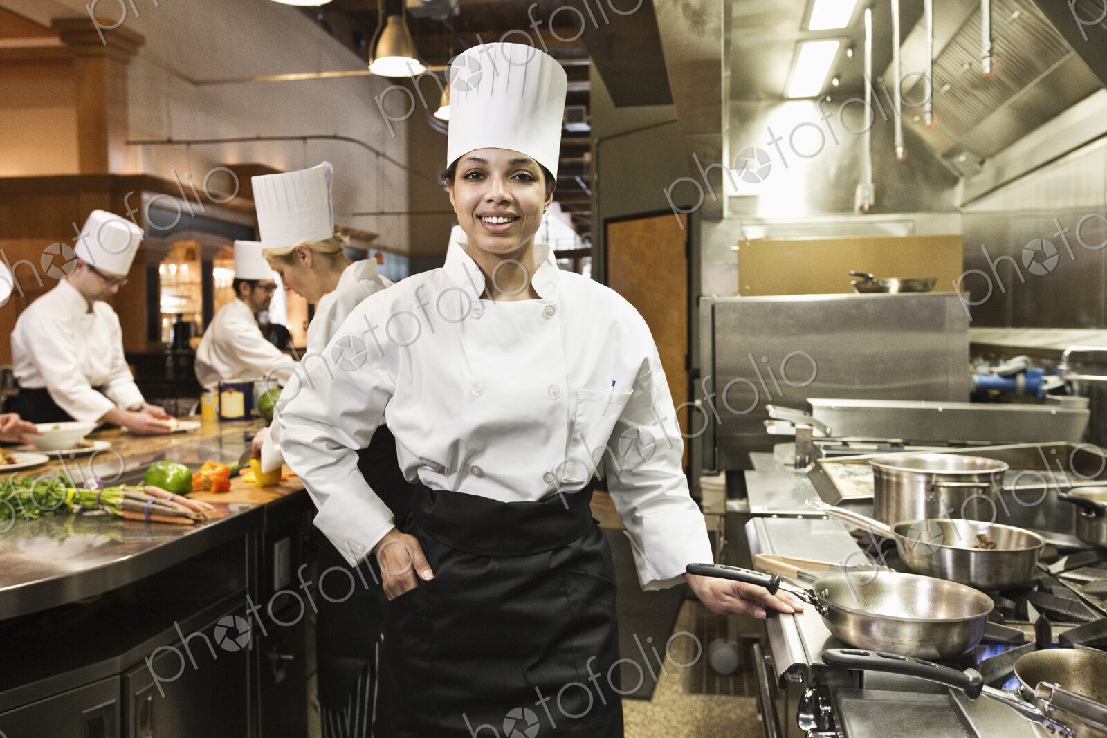 A portrait of a young black female chef in a commercial kitchen with her crew working…