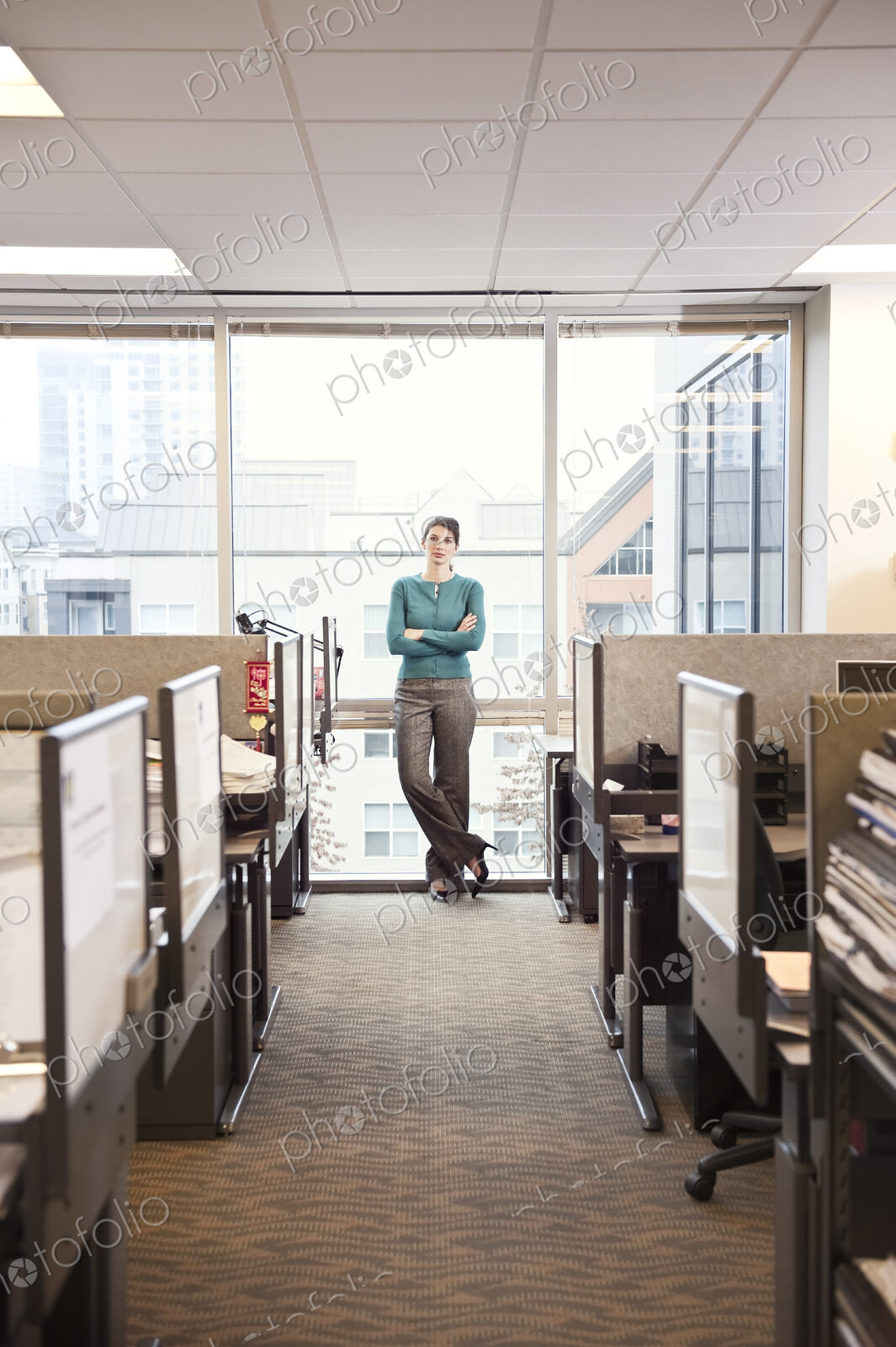 A portrait of a young businesswoman standing at a window near cubicles in a corporate…