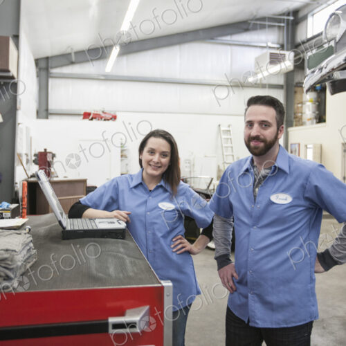 A portrait of two caucasian male and female mechanics in an antique car repair shop.
