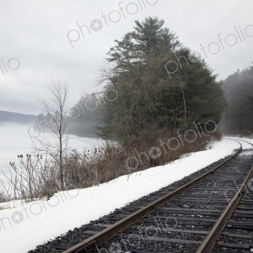 A railway track, train tracks in the snow, by a lake in the Vermont landscape, mist…