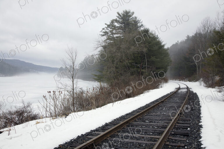 A railway track, train tracks in the snow, by a lake in the Vermont landscape, mist…