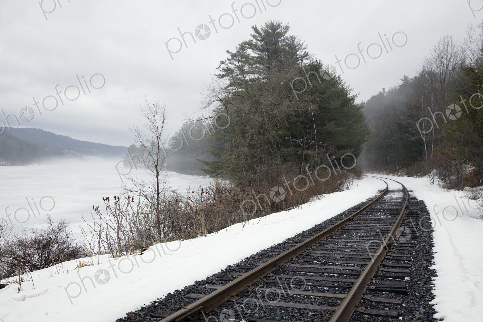 A railway track, train tracks in the snow, by a lake in the Vermont landscape, mist…