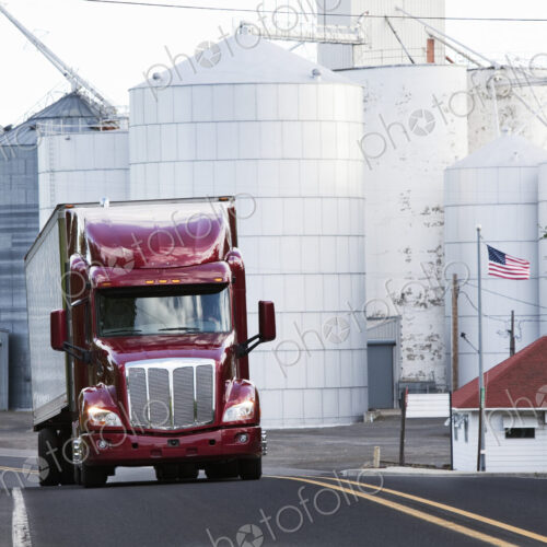 A red commercial truck driving past grain elevators in the farm country of eastern…