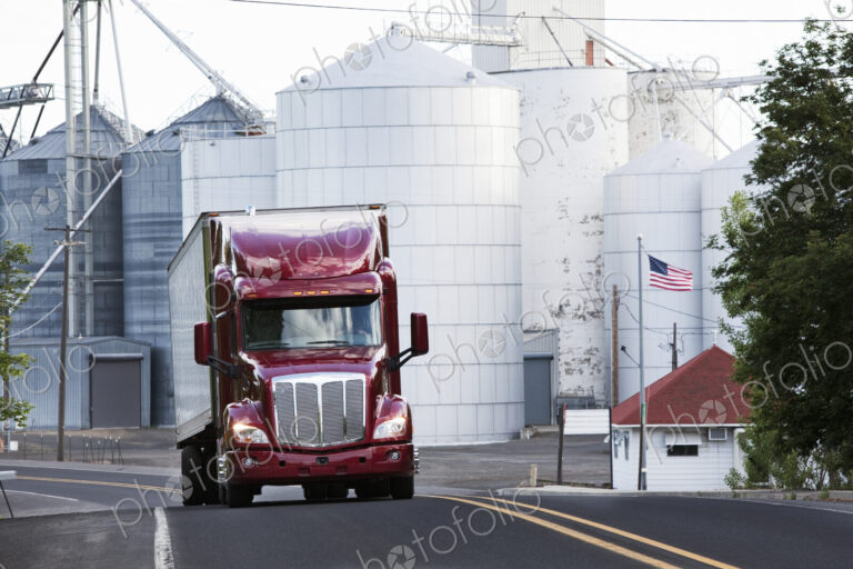 A red commercial truck driving past grain elevators in the farm country of eastern…